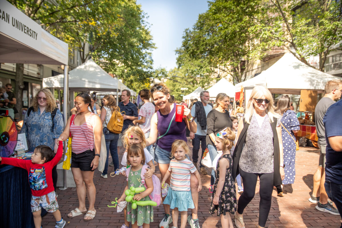 a photo of people during Oaktoberfest on a sunny day walking, smiling and buying items on tents