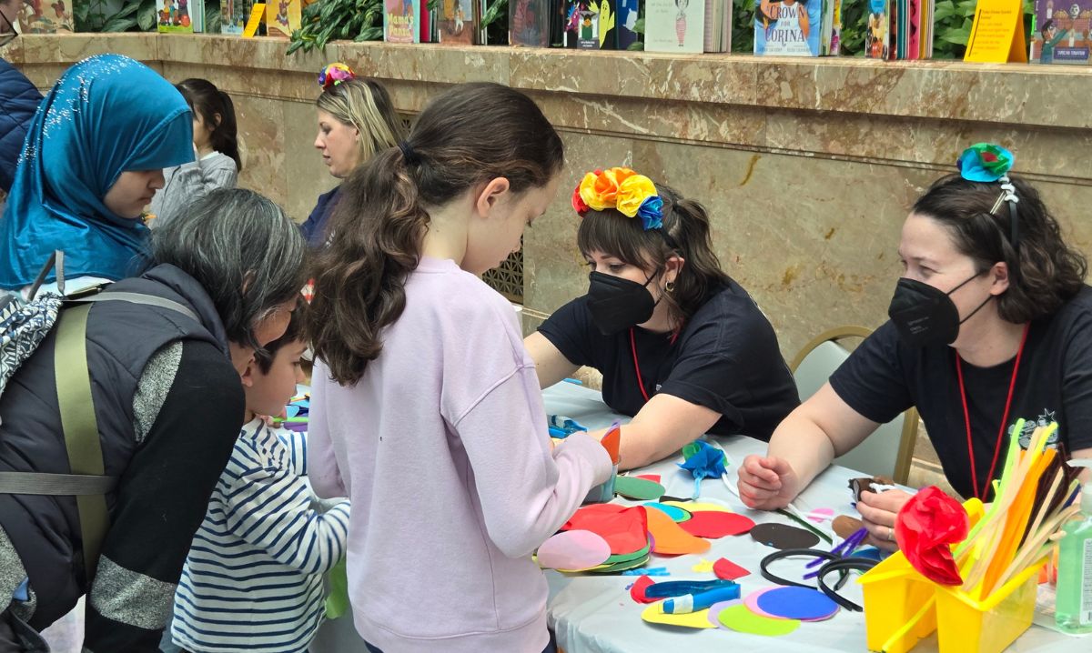 Families participating in a Mardi Gras-themed craft activity at the Chicago Public Library, with children and adults creating colorful paper decorations under the guidance of staff members wearing festive flower crowns.
