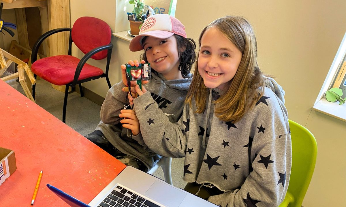 Two smiling students showcasing a small electronic programming project with an LED display, sitting at a table with a laptop during a hands-on STEM activity.
