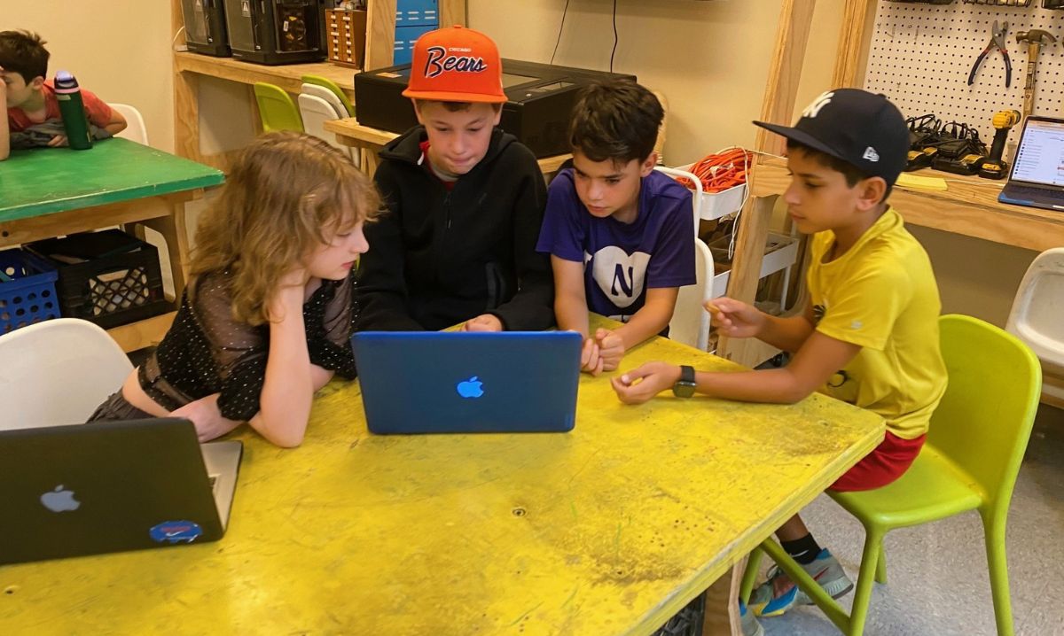 Four students collaborating around a laptop in a hands-on classroom environment, discussing ideas and working together on a project.