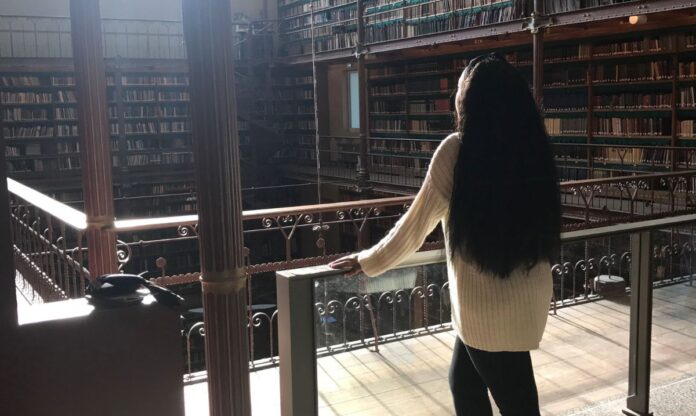 A woman standing on the balcony of a historic reading room at the Chicago Public Library, surrounded by shelves filled with countless books and basking in warm natural light.