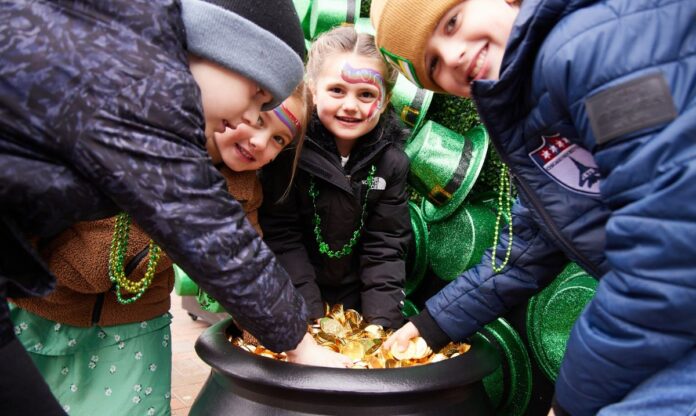 Children excitedly reach into a pot of gold coins at the St. Patrick’s Day celebration at Gallagher Way in Chicago. They are dressed in festive attire, including green bead necklaces and face paint, as they pose in front of a backdrop decorated with green leprechaun hats. The joyful expressions capture the spirit of the holiday celebration.