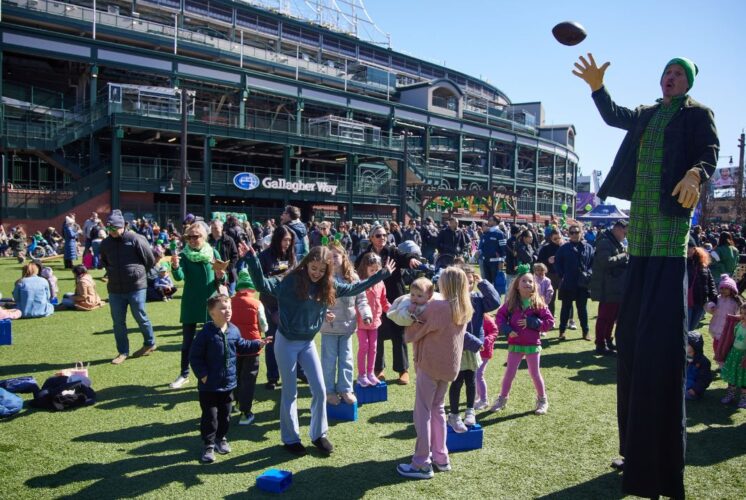 Families and children celebrate St. Patrick’s Day at Gallagher Way in Chicago with outdoor games and entertainment. A performer on stilts, dressed in green plaid, tosses a football while kids and parents participate in activities on the artificial turf. The festive crowd, dressed in green, enjoys the sunny day with Wrigley Field as the backdrop. The lively atmosphere includes games, dancing, and holiday-themed fun.