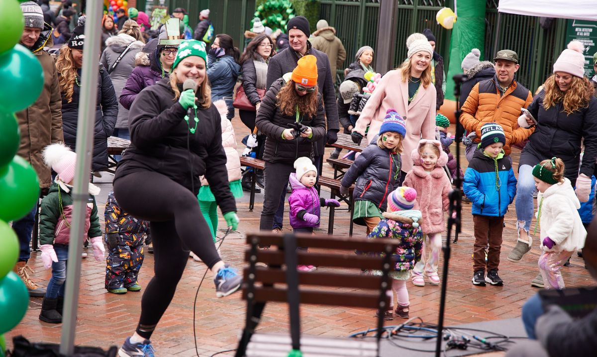 Families and children celebrate St. Patrick's Day at Gallagher Way in Chicago with a lively dance party. A woman in festive green attire leads the crowd in dancing, as kids and parents join in the fun. Attendees, dressed in warm coats and hats, enjoy the energetic atmosphere with green decorations, balloons, and festive accessories. The background features a crowd of families, benches, and the entrance to Wrigley Field.